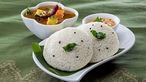 Two white rice cakes on a green leaf, accompanied by a bowl of soup and a small bowl of chutney. - Vrindavan Greens Resort (Udaipur)