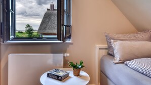 Ein Schlafzimmer mit Fensterblick auf ein Haus, einem Heizkörper, einem Nachttisch mit Buch und einer Pflanze.