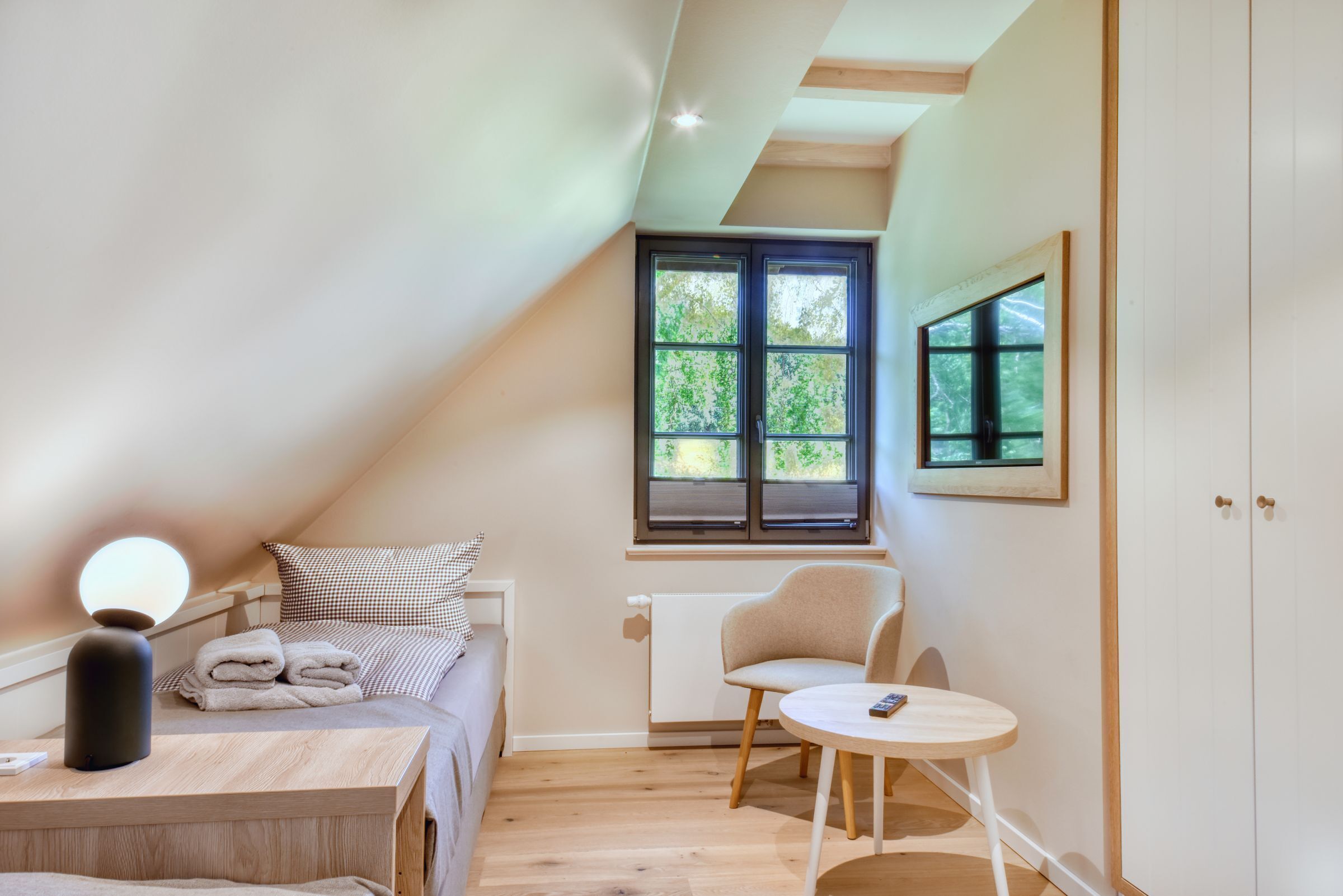 Un dormitorio con cama, una mesita de noche con lámpara, una silla, una pequeña mesa y una ventana con vista a árboles.