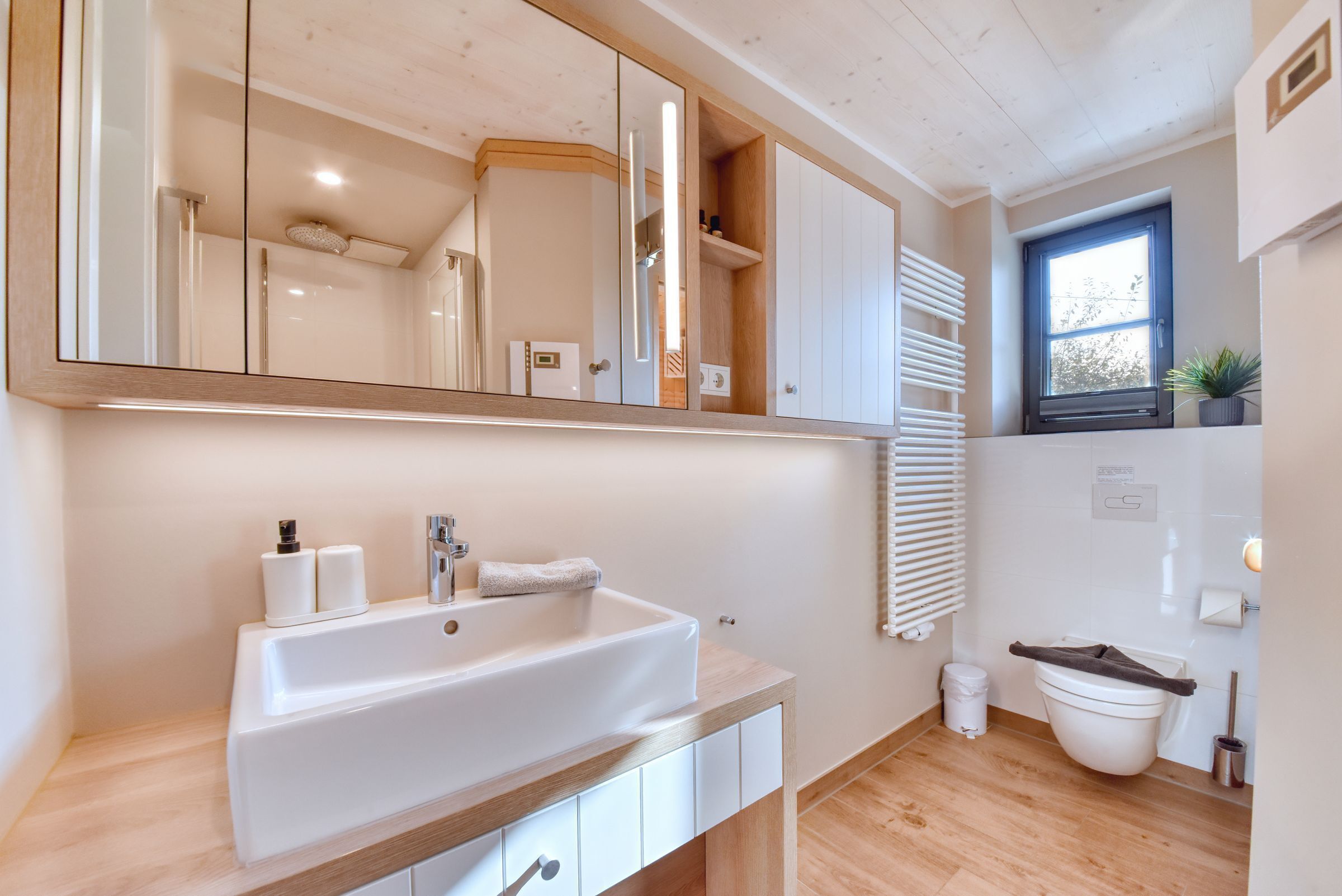 Une salle de bain moderne avec un grand miroir, un lavabo blanc, un meuble-lavabo en bois et une fenêtre avec des stores.