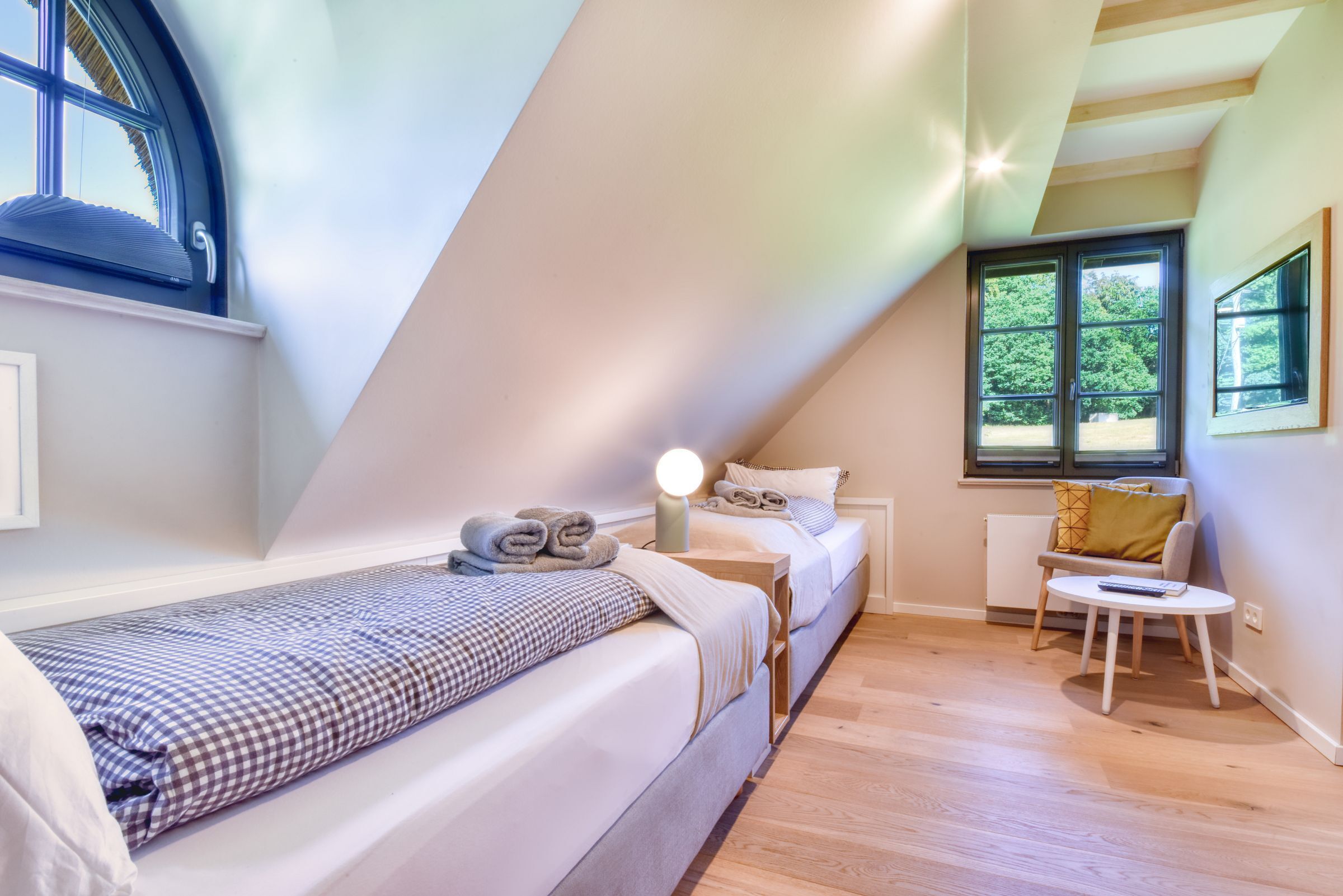 A bedroom with a sloped ceiling, a bed, a bedside table with a lamp, a small table with a chair, and a window with a view of greenery.