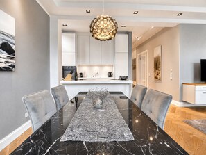 A modern dining area with a marble table, grey upholstered chairs, a decorative centerpiece, and a kitchen in the background.