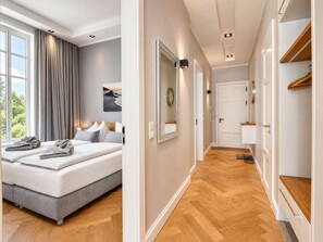 Hotellihuone, jossa on sänky, näkymä ulos, käytävä, joka johtaa ovelle, ja puulattia.