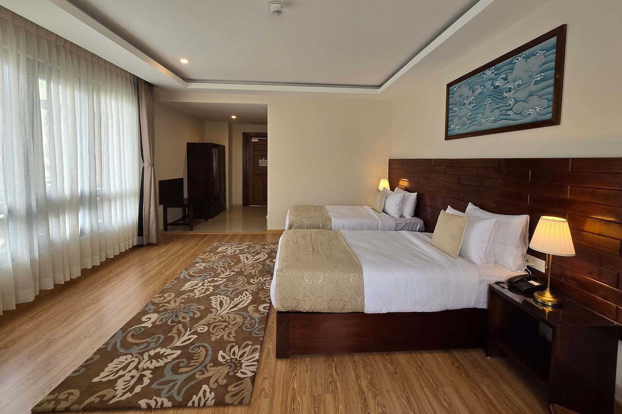 Habitación de hotel con dos camas, cabecera de madera, mesita de noche con lámpara, ventana grande con cortinas traslúcidas y un cuadro en la pared.