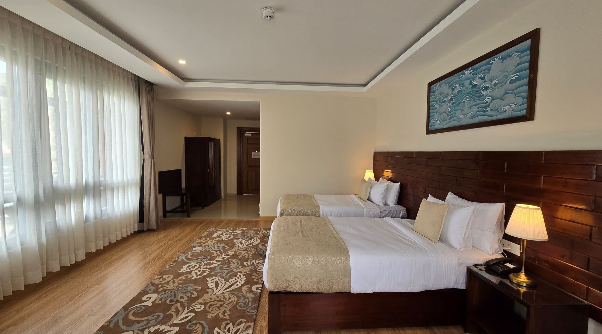 A hotel room with two beds, a wooden headboard, a large window with sheer curtains, a patterned rug, and a painting on the wall.