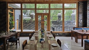 Un comedor con una larga mesa de madera dispuesta para una comida, ventanas amplias que dan a un muro de piedra y follaje, y una chimenea de piedra.
