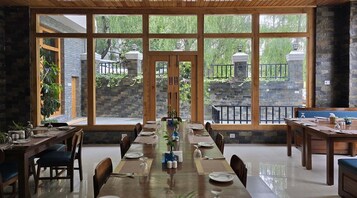 Un comedor con una larga mesa de madera dispuesta para una comida, ventanas amplias que dan a un muro de piedra y follaje, y una chimenea de piedra.