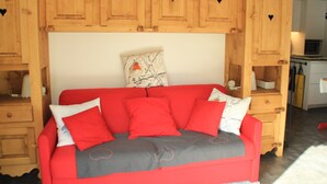 A red sofa with grey and white cushions, wooden cabinets with heart-shaped cutouts, and a kitchen area in the background. - Moulin Ml161 Telecabine & Nature 6 Pers (Chatel)