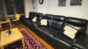 Ein Wohnzimmer mit einer schwarzen Ledersofa, einem Holztisch, einem gemusterten Teppich, einer kleinen Pflanze und einer Wanduhr.