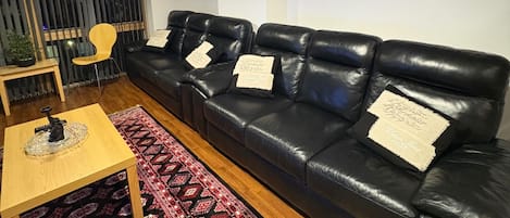 En stue med en sort lædersofa, et træbord, et mønstret tæppe, en lille plante og et vægur.