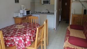 A compact kitchen with a microwave, toaster, and coffee maker. There is a dining table with a red tablecloth and a wooden chair. A small kitchenette is visible in the background. - Christina CR8 Family & Mountain 6 Pers (Chatel)