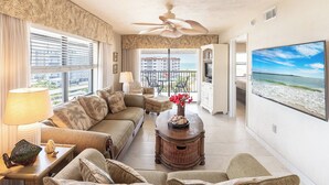 A living room with a sofa, a coffee table with a vase, a TV, and a balcony with a view of the ocean.
