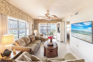 A living room with a sofa, a coffee table with a vase, a TV, and a balcony with a view of the ocean.