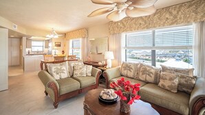 A living room with a sofa, a coffee table with decorative items, a ceiling fan, and a view through large windows.