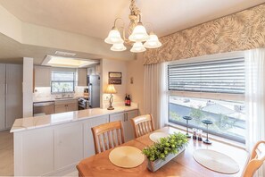 A modern kitchen with a central island, a dining area with a wooden table, and a view through large windows.