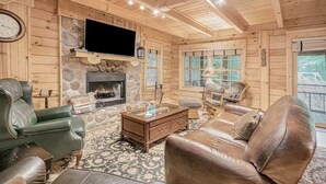 A cozy log cabin interior with a stone fireplace, leather furniture, and a flat-screen TV mounted above it. - Cozy Cabin Getaway with Private Hot Tub & Lake Access in Saugatuck! (Saugatuck)