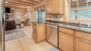 A log cabin interior with wooden floors, a stone fireplace, a kitchen with granite countertops and wooden cabinets, and a large window offering a view of the outdoors. - Cozy Cabin Getaway with Private Hot Tub & Lake Access in Saugatuck! (Saugatuck)