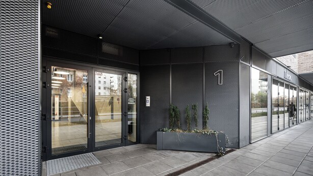 Una entrada moderna de edificio con puertas de vidrio, un muro de malla metálica y una jardinera con follaje.