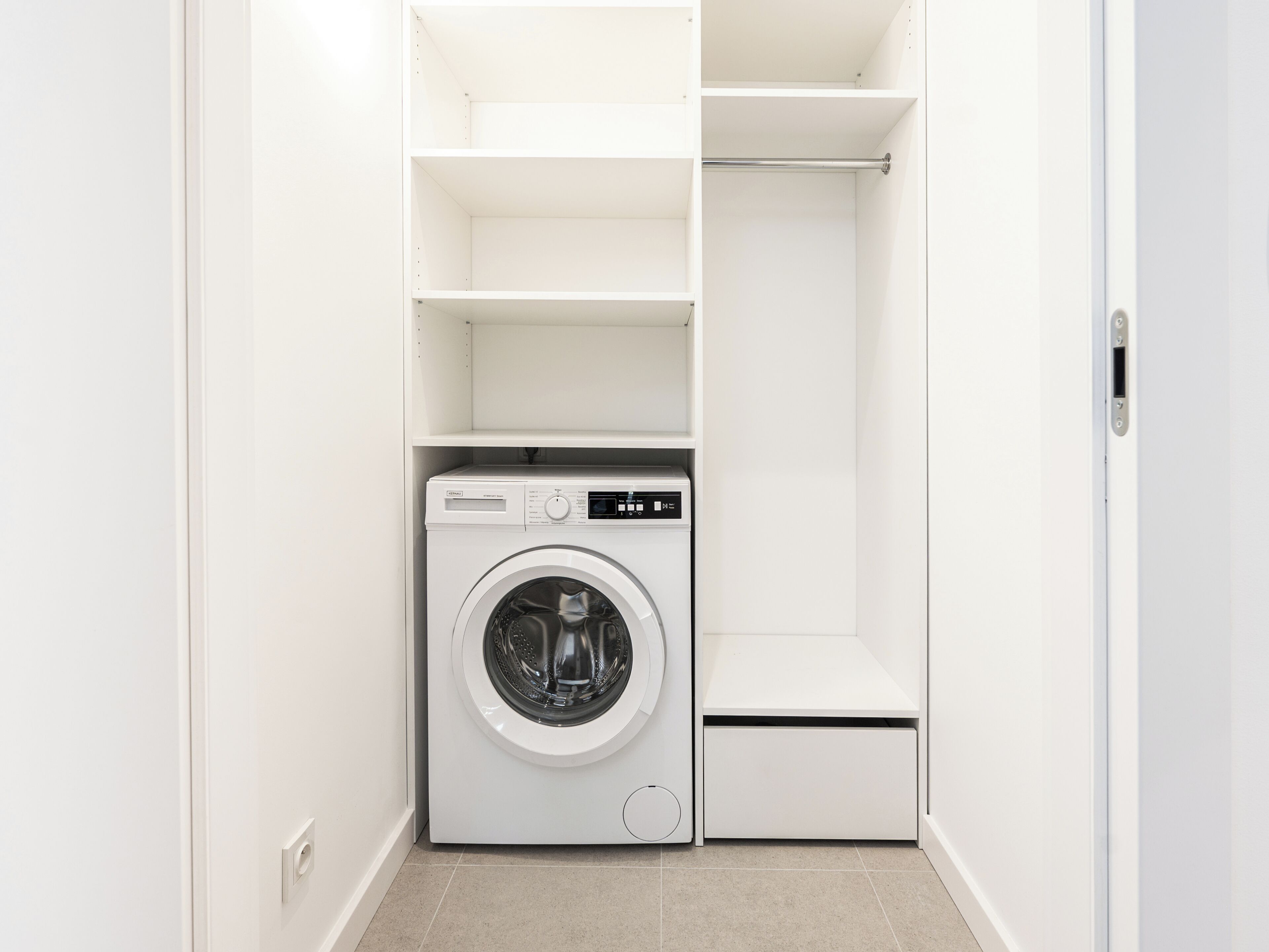 Une petite buanderie équipée d’un lave-linge à chargement frontal, d’étagères intégrées et d’un petit meuble de rangement.