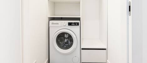 Une petite buanderie équipée d’un lave-linge à chargement frontal, d’étagères intégrées et d’un petit meuble de rangement.