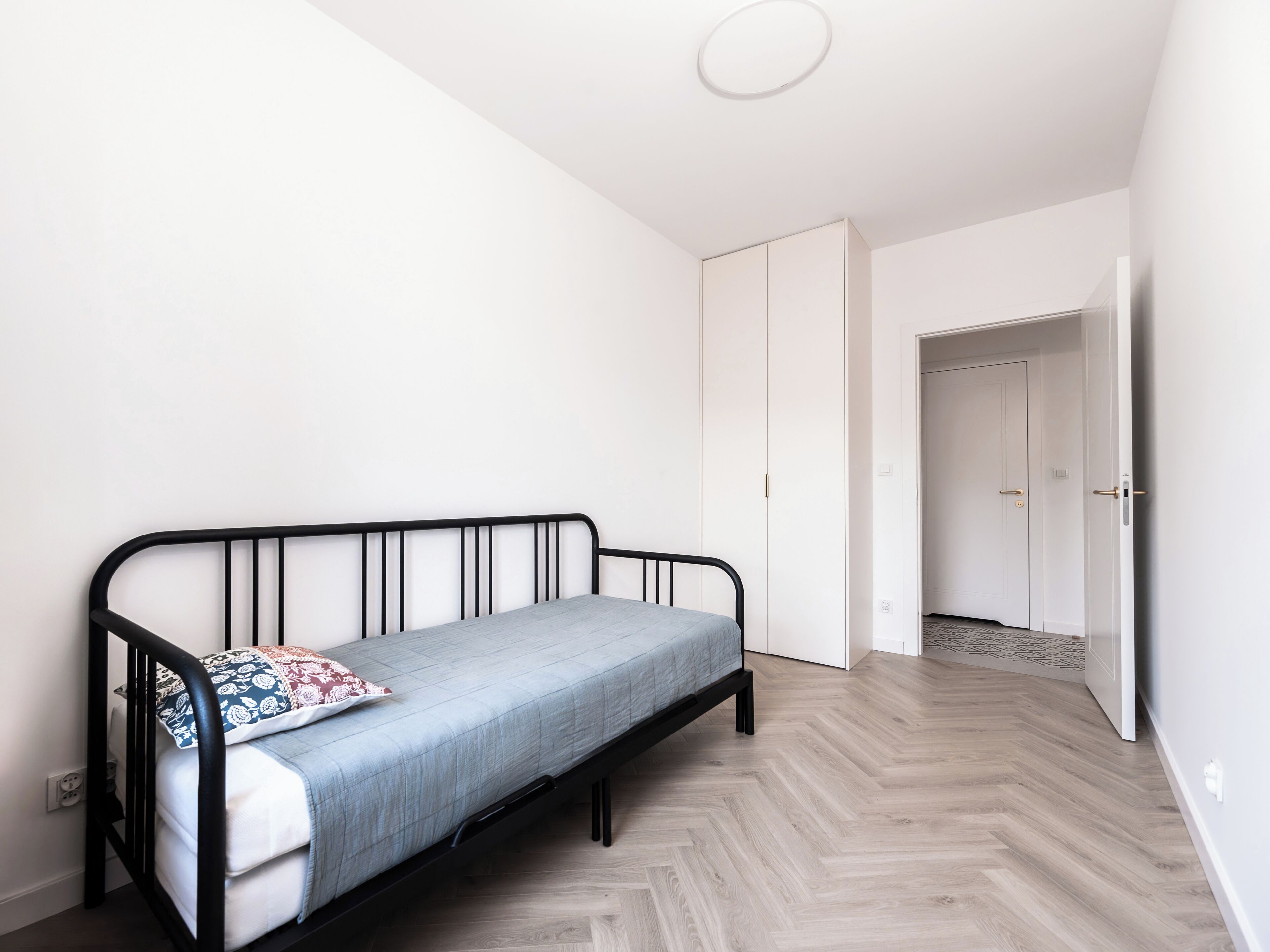 Une petite chambre avec un lit simple, un cadre en métal, un matelas bleu clair et un oreiller à motifs. La chambre a un parquet en bois disposé en chevrons et des murs blancs. On y trouve une armoire blanche et une porte qui donne sur une autre pièce.