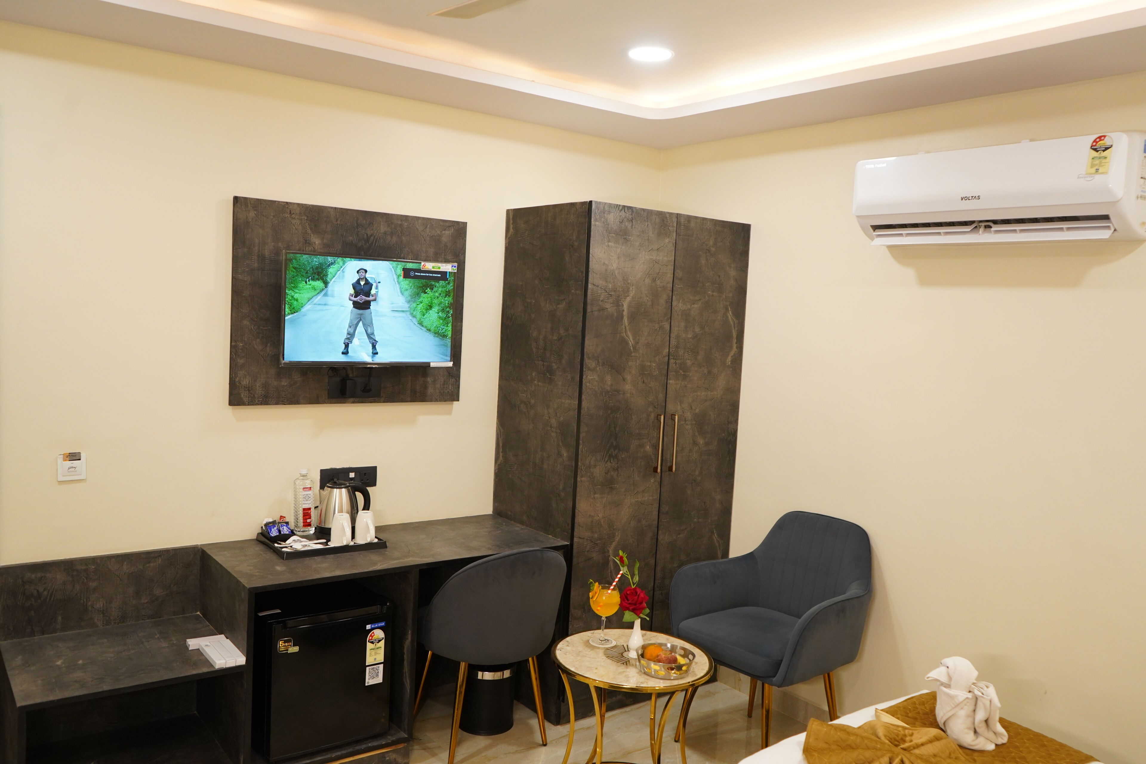 Una habitación de hotel moderna con un televisor de pantalla plana montado en la pared, un armario de madera oscura, una mesita redonda con un jarrón de flores y una zona de estar con un sillón azul.