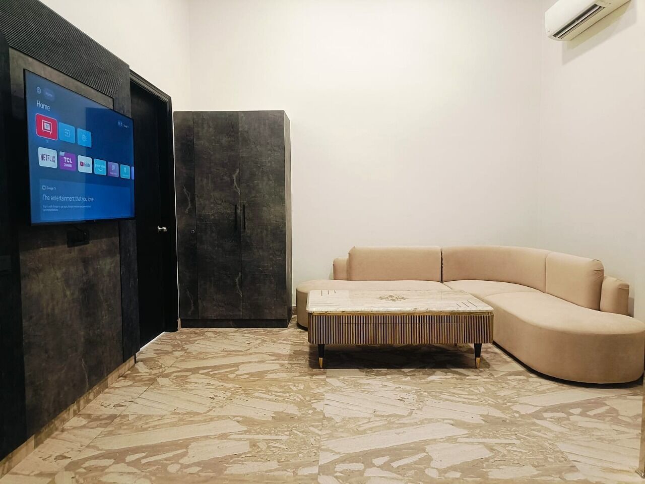 Un soggiorno moderno con una TV a schermo piatto montata su una parete scura, un divano componibile beige e un poggiapiedi decorato.