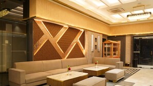 A modern lobby with a beige sofa, two matching ottomans, and a coffee table with a geometric patterned wall behind it. - Shrigo Hotels Banaras (Varanasi)