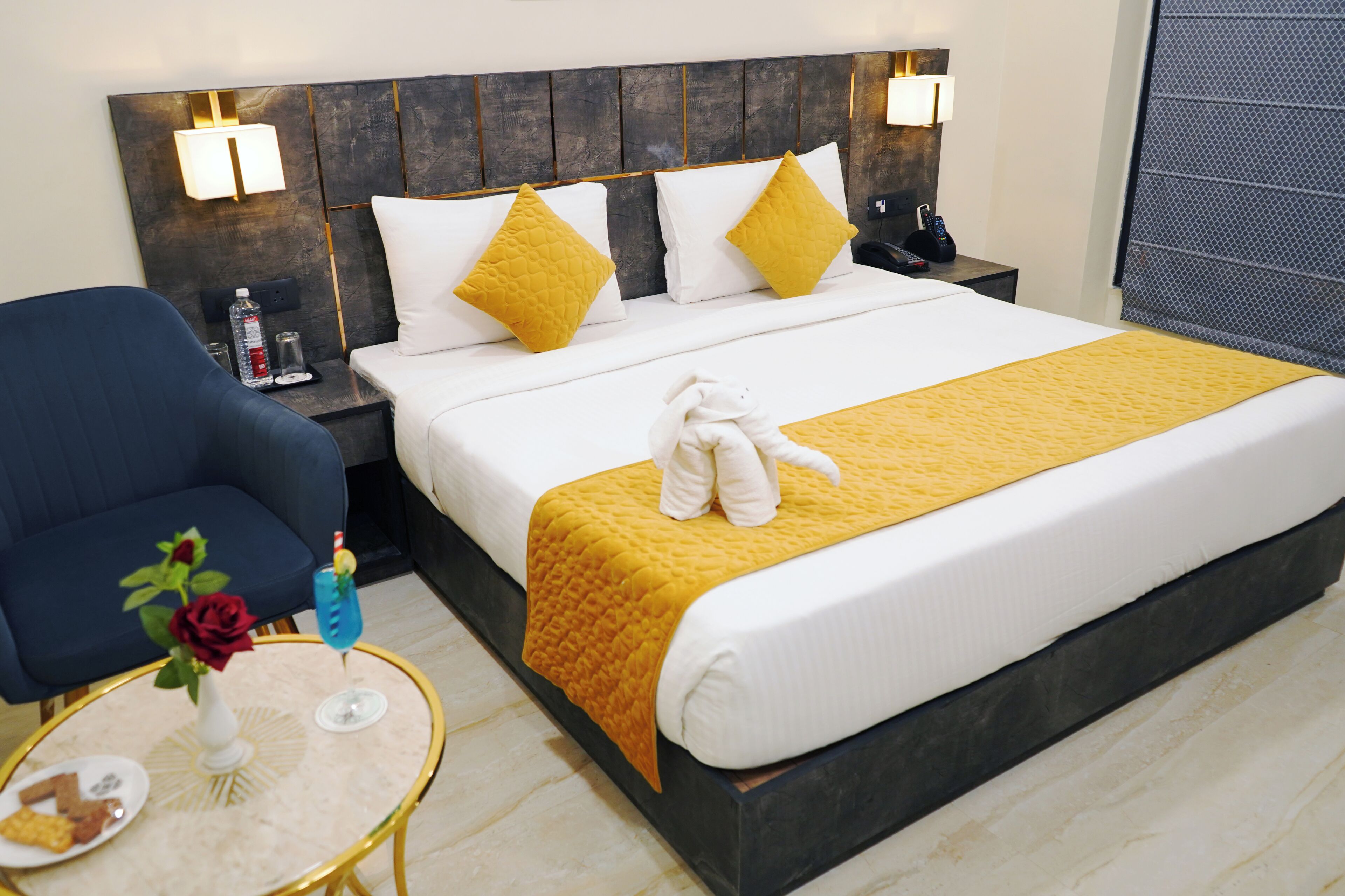 Une chambre d’hôtel avec un grand lit, une table de chevet avec une lampe, une chaise et une petite table ronde ornée d’un vase et d’une bouteille.
