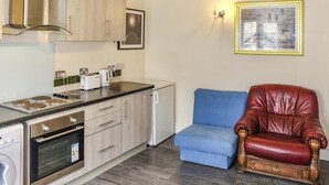 A compact kitchen with a built-in oven, a coffee maker, and a toaster. There is a blue chair and a red leather armchair. - Host Stay Apartment 4 Church Walk (Melksham)