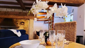 A dining table set with white plates, bowls, cutlery, and crystal glasses, surrounded by a woven chair and a blue sofa in a rustic room with exposed beams and a stone wall. - Host Stay Gwenelin Cottage (Evesham)