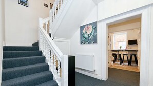Un escalier avec des marches recouvertes de moquette grise et des balustrades blanches, menant à une pièce où se trouvent un radiateur, un tableau et un coin cuisine avec des tabourets.