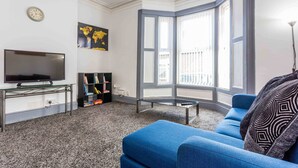 A living room with a blue sofa, a flat-screen TV, a bookshelf, and a large window with a view. - Host Stay Bambro Apartment (Sunderland)