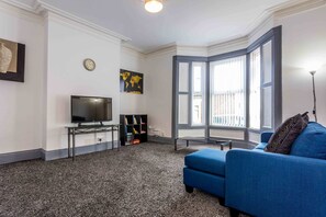 A living room with a blue sofa, a flat-screen TV, a bookshelf, and a large window with blinds. - Host Stay Bambro Apartment (Sunderland)
