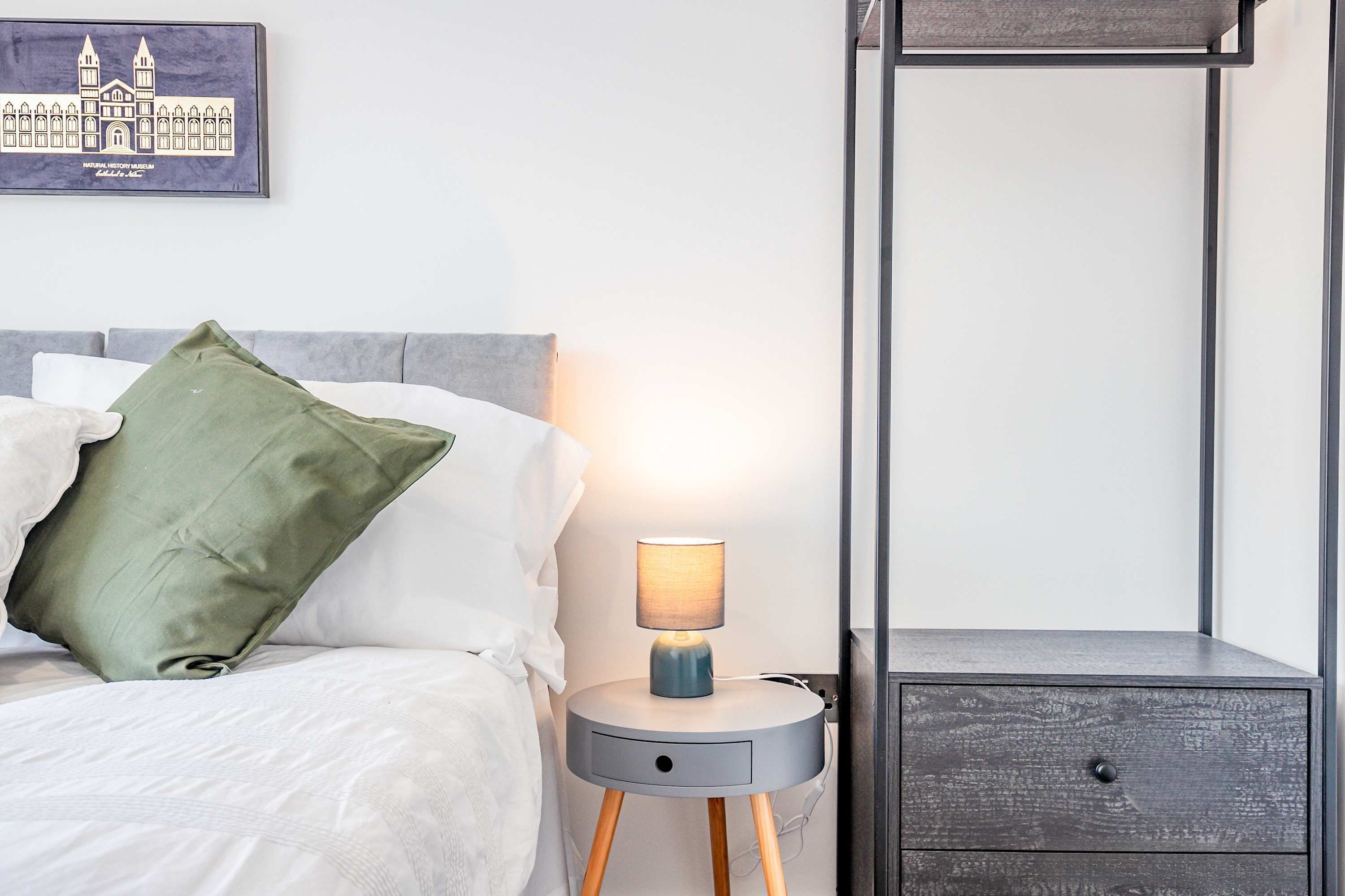 A neatly made bed with a green pillow, a bedside table with a lamp, and a small drawer unit.