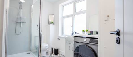 A modern bathroom with a glass-enclosed shower, a toilet, a washing machine, and a window providing natural light.