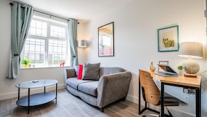 A modern living room with a grey sofa, a round glass table, a wooden desk with a laptop, and a lamp. - Host Stay Croydon Road Apartment (Caterham)