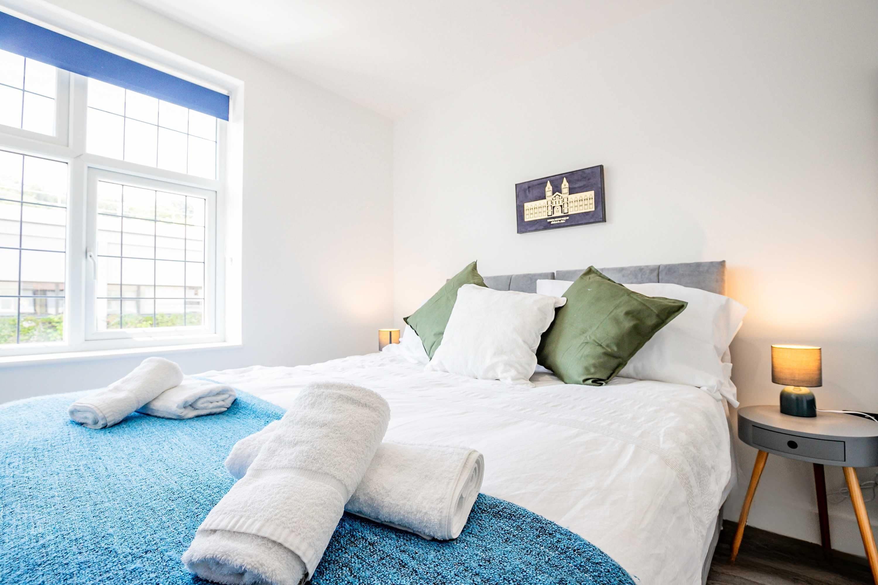 Een net opgemaakt bed met witte beddengoed en groene kussens, een blauwe plaid, en opgerolde witte handdoeken op een blauw vloerkleed.