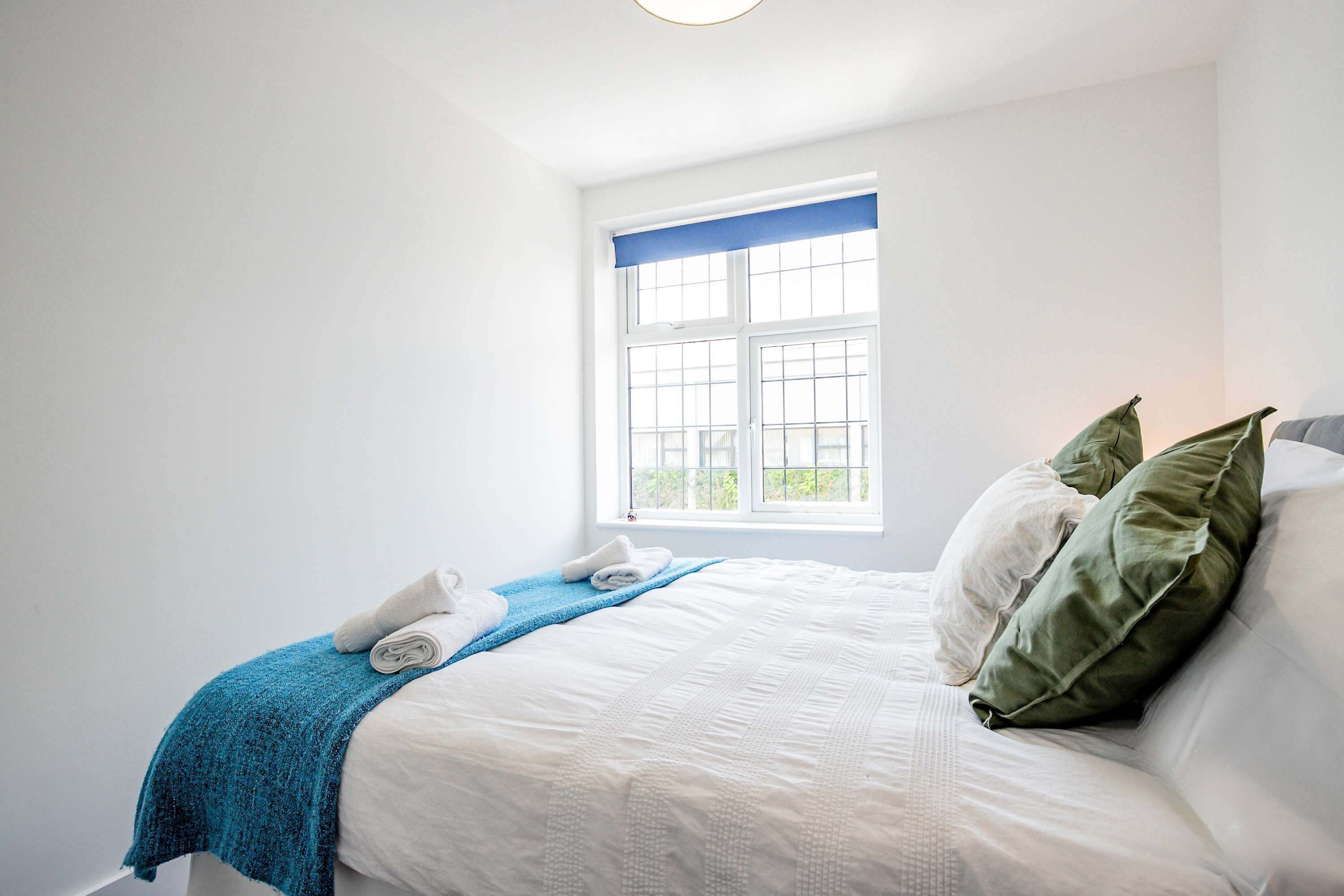 A neatly made bed with a blue blanket and pillows, a window with blinds, and a white wall.