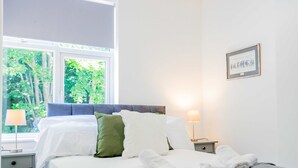 A hotel room with a bed, two bedside lamps, a framed picture on the wall, and a window with a view of trees. - Host Stay Croydon Road Dwelling (Caterham)