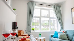 A cozy living room with a sofa, a table with food and drinks, a window with curtains, and a small plant on the wall. - Host Stay Croydon Road Dwelling (Caterham)