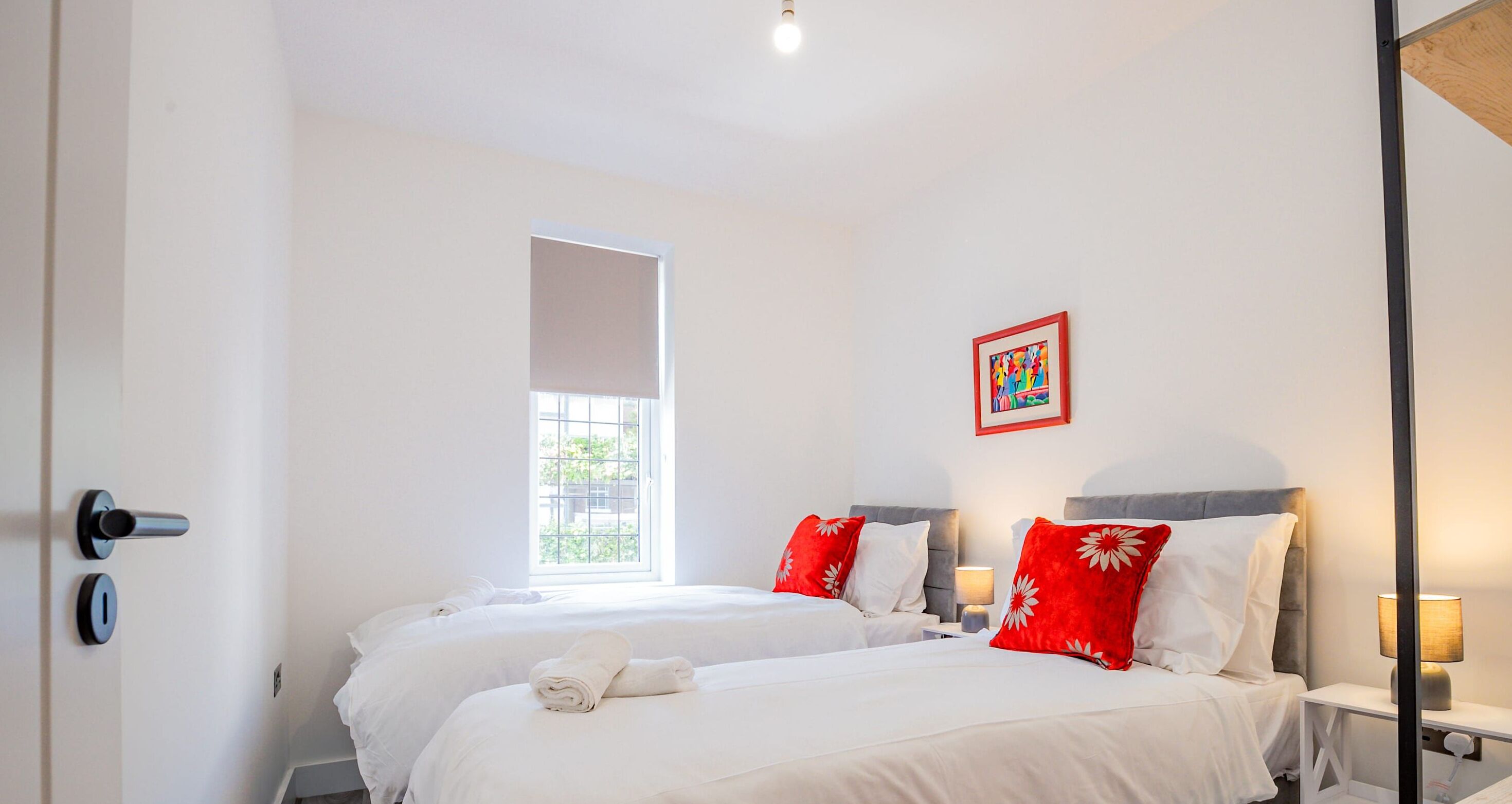 A hotel room with two beds, a window, a painting, and a nightstand with a lamp.
