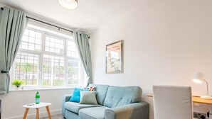 A room with a light blue sofa, a small white table, a lamp, and a window with curtains. - Host Stay Croydon Road Dwelling (Caterham)