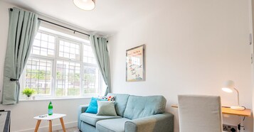 A room with a light blue sofa, a small white table, a lamp, and a window with curtains.