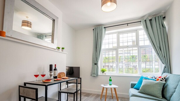 A modern living room with a dining area, a sofa, a TV, and a window with curtains. - Host Stay Croydon Road Dwelling (Caterham)