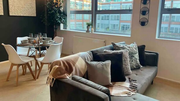 A modern living room with a grey sofa, white chairs, and a dining table set for four. Large windows offer a view of buildings outside. - Host Stay Great Hampton Apartment (Birmingham)