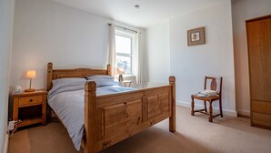 A bedroom with a wooden bed, a nightstand with a lamp, a chair, a window with curtains, and a radiator. - Host Stay Wellington Apartment (Horsham)