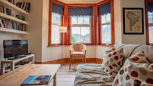 A cozy room with a wooden floor, a bookshelf, a television, a wicker chair, a sofa with polka dot cushions, and a large window with curtains. - Host Stay Wellington Apartment (Horsham)