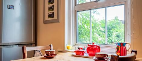 A dining area with a wooden table, chairs, a refrigerator, and a window with a view of greenery.