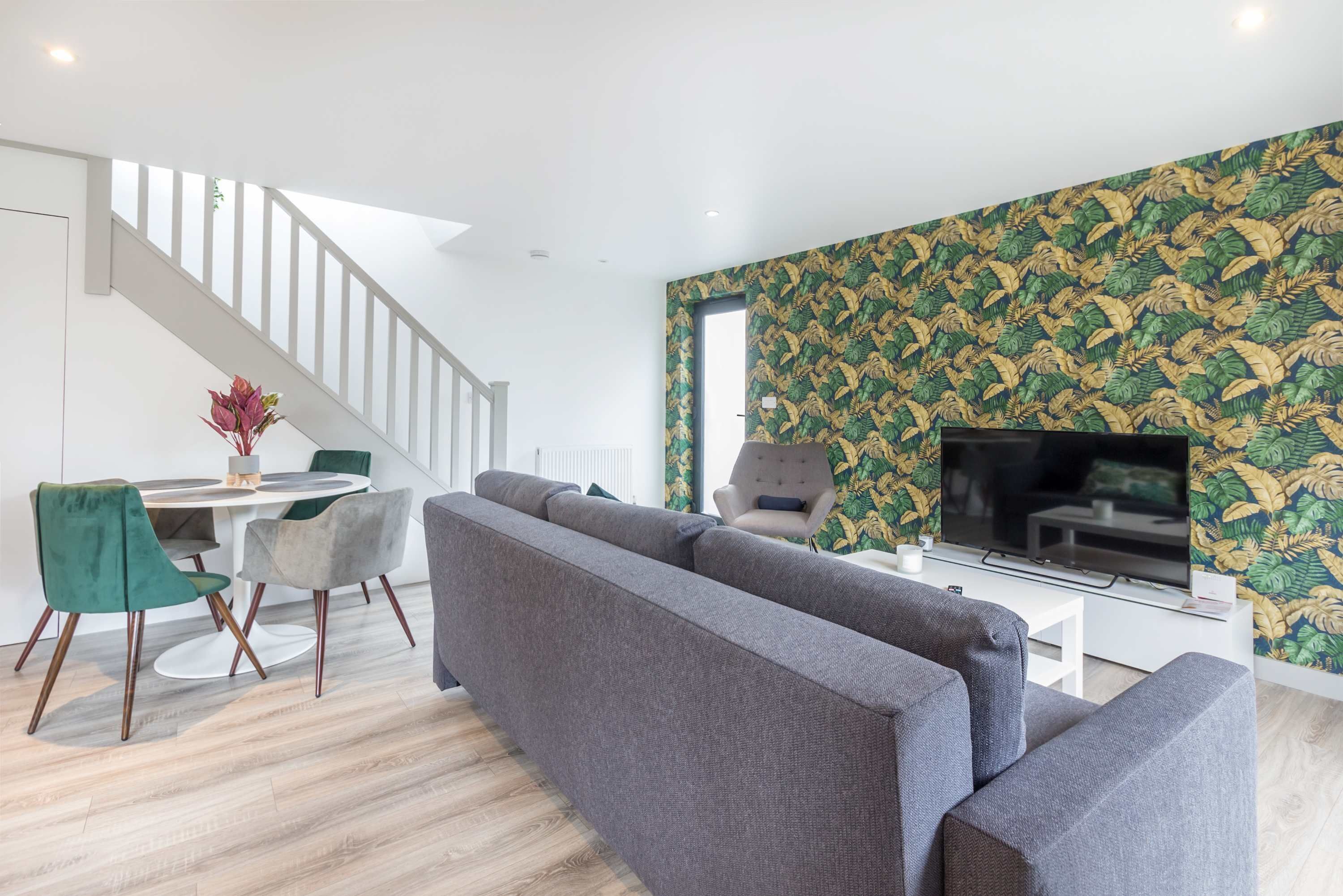 Ein modernes Wohnzimmer mit einem grauen Ecksofa, einem Esstisch mit Stühlen, einer Treppe und einem Fernseher auf einem weißen Lowboard.